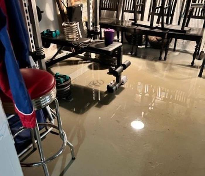 Standing water in a basement with put-up tables, chairs, and exercise equipment on a weight bench