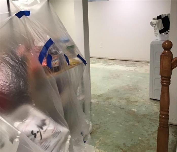 A demolished basement floor with removed baseboards and bagged contents near a banister leading upstairs